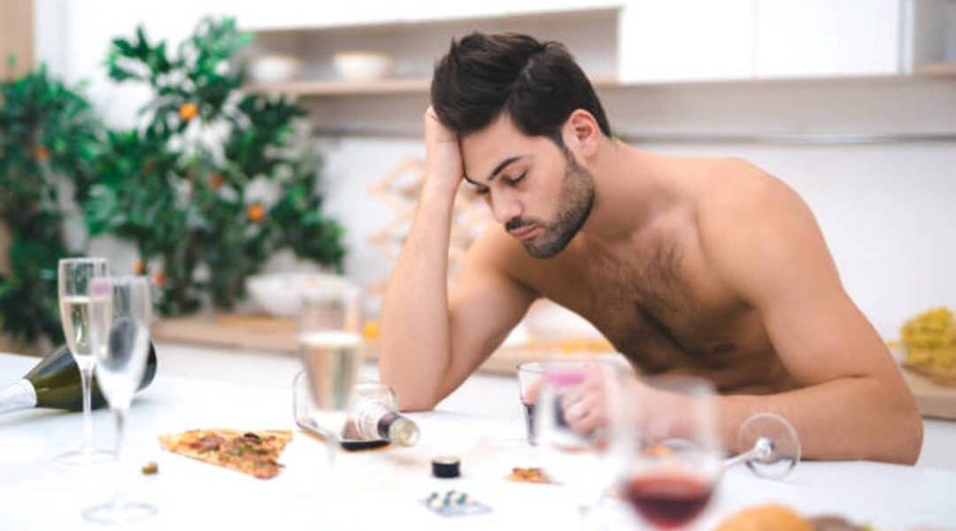 A shirtless man with dark hair is sitting at a table with his elbow resting on the surface and his head in his hand, appearing distressed or fatigued. The table is cluttered with glasses, a wine bottle, and remnants of food, including a slice of pizza. Perhaps he’s contemplating the benefits of convenient IV treatments.