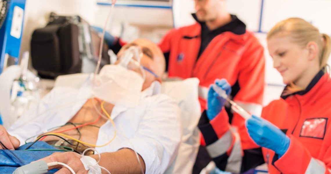 A patient is lying on a hospital bed with an oxygen mask and medical monitoring equipment attached. A healthcare professional in an orange jacket is adjusting the equipment, while another in an orange jacket prepares a syringe. Both certified IV nurses are wearing blue gloves, ensuring safe and effective IV therapy.