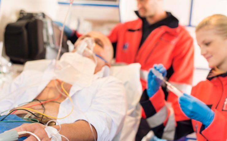 A patient is lying on a hospital bed with an oxygen mask and medical monitoring equipment attached. A healthcare professional in an orange jacket is adjusting the equipment, while another in an orange jacket prepares a syringe. Both certified IV nurses are wearing blue gloves, ensuring safe and effective IV therapy.