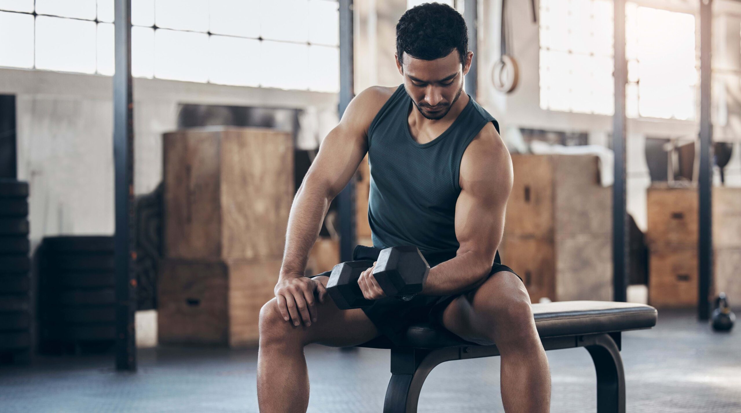 A person wearing a sleeveless shirt and shorts is seated on a bench at a gym, concentrating while performing a bicep curl with a dumbbell. In the background, there are large wooden boxes, gym equipment, and windows letting in natural light.