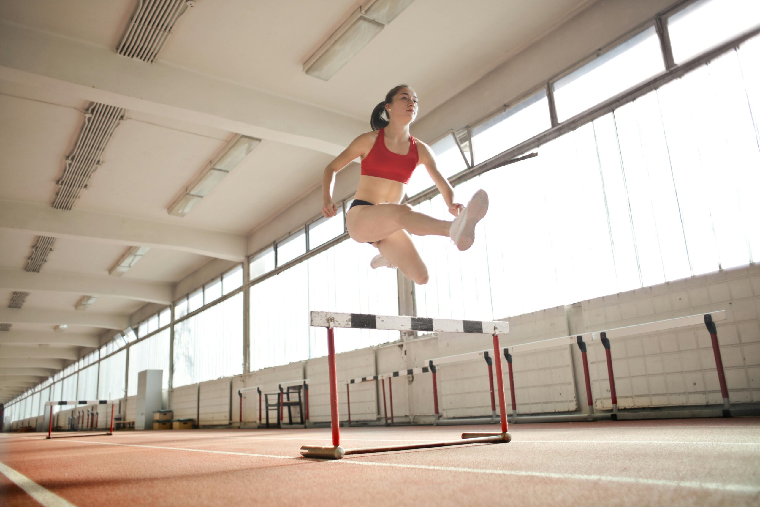 A female athlete in a red sports bra and black shorts is airborne, leaping over a hurdle on an indoor track. The spacious facility with large windows lets in natural light, providing an ideal setting for her rigorous training and subsequent recovery, including the benefits of recovery IV drips for women.