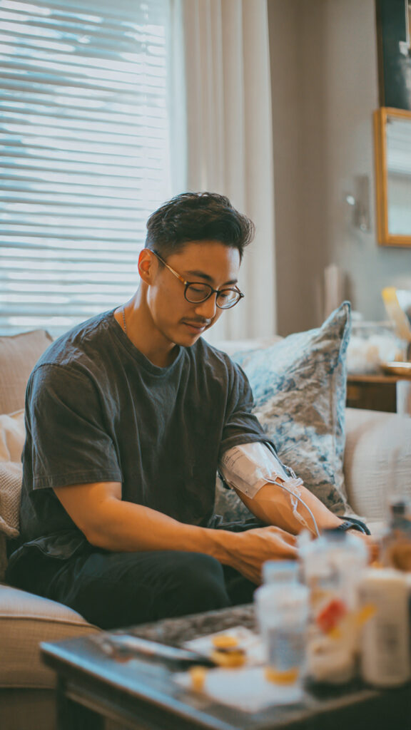 A man with glasses sits on a couch, receiving an IV drip in his arm. He looks down, appearing calm and relaxed. There are bottles and items on a table in front of him, and a window with blinds in the background.