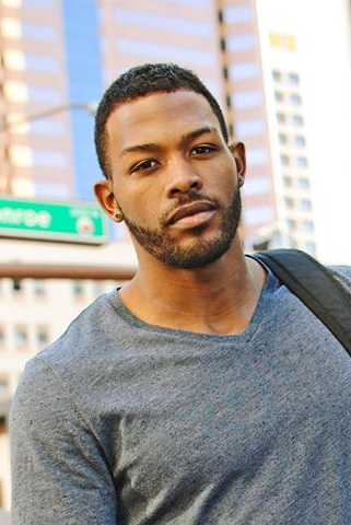 A man wearing a gray t-shirt and jeans stands outdoors in an urban area, holding a black jacket and bag. Tall buildings and a "Monroe" street sign are visible in the background.