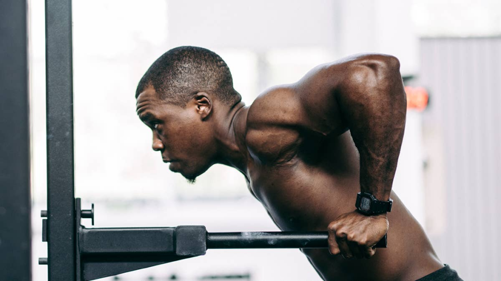A man wearing a smartwatch performs a dip exercise on parallel bars, showing strong arm and shoulder muscles, in a brightly lit gym setting.
